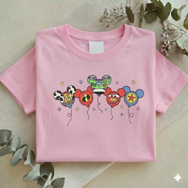 Może przedstawiać: Jasnor&oacute;żowa koszulka z kolorową grafiką balon&oacute;w z postaciami z kresk&oacute;wek. Balony są zaprojektowane tak, aby przypominały Myszka Miki, Buzz Lightyear i Zygzaka McQueena. Projekt zawiera gwiazdki i sznurki od balon&oacute;w.