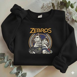 Może przedstawiać: Biały ceramiczny kubek z czarnym uchwytem i napisem "But First, Coffee" czarną, pogrubioną czcionką. Kubek stoi na białej powierzchni. Kubek ma około 10 cm wysokości.