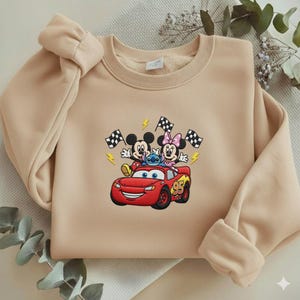 May include: Beige sweatshirt featuring an embroidered design of Mickey Mouse, Minnie Mouse, and Stitch riding in a red Lightning McQueen race car. The design includes checkered flags and lightning bolts. The sweatshirt has a crew neck and long sleeves.
