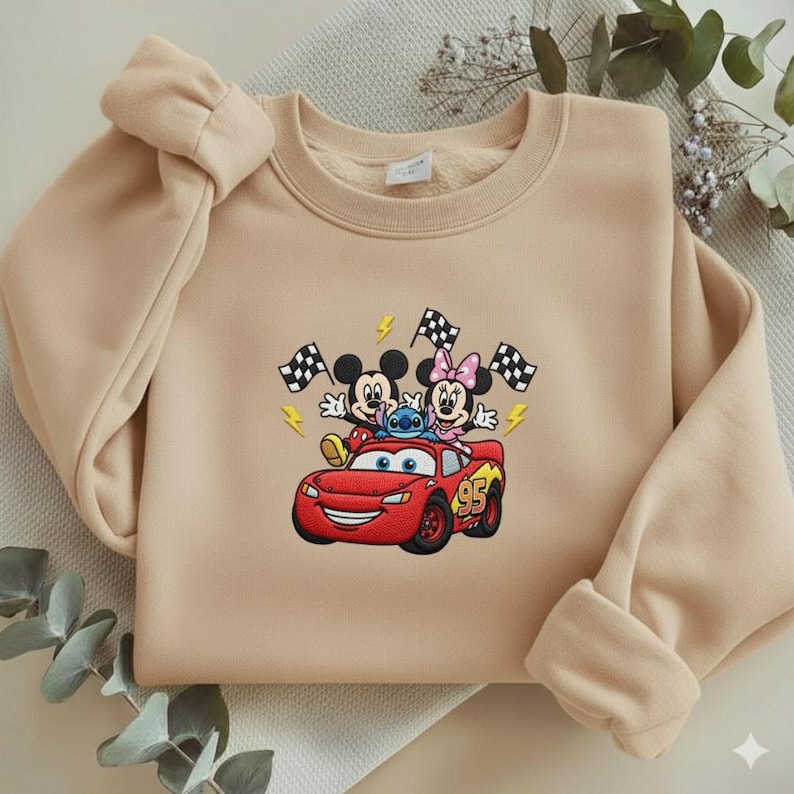 May include: Beige sweatshirt featuring an embroidered design of Mickey Mouse, Minnie Mouse, and Stitch riding in a red Lightning McQueen race car. The design includes checkered flags and lightning bolts. The sweatshirt has a crew neck and long sleeves.