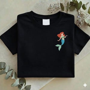 May include: Black t-shirt featuring a colorful illustration of a mermaid with red hair, a green top, and a blue tail. The shirt is folded neatly, with a blank white tag visible near the neckline. The design is on the left side of the shirt.