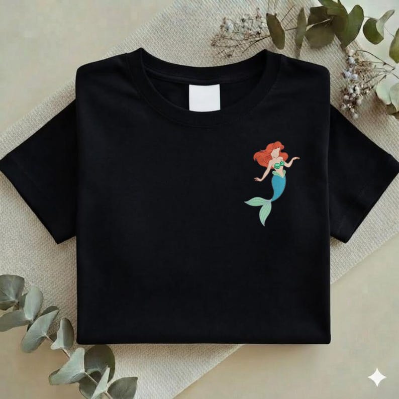 May include: Black t-shirt featuring a colorful illustration of a mermaid with red hair, a green top, and a blue tail. The shirt is folded neatly, with a blank white tag visible near the neckline. The design is on the left side of the shirt.