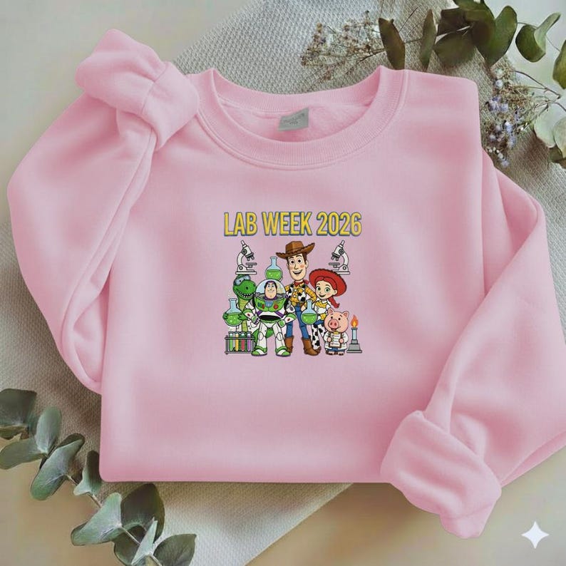 Puede incluir: Sudadera rosa claro con el texto "LAB WEEK 2026" en amarillo. El dise&ntilde;o presenta personajes de Toy Story, como Woody y Buzz Lightyear, junto con equipos de laboratorio.