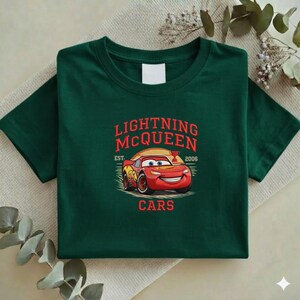 Puede incluir: Camiseta verde oscuro con un dise&ntilde;o de Rayo McQueen de la pel&iacute;cula Cars. El dise&ntilde;o incluye el texto "LIGHTNING McQUEEN" en rojo, con "EST. 2006" y la palabra "CARS" debajo del coche.