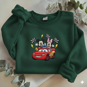 May include: A dark green sweatshirt featuring an embroidered design of Mickey Mouse, Minnie Mouse, Stitch, and Lightning McQueen. The design includes checkered flags and lightning bolts. The number 95 is on the car.