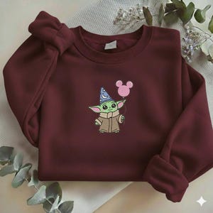 Op de afbeelding: Bordeaux sweatshirt met een geborduurd stripfiguur met een tovenaars hoed en een roze ballon. Het personage is groen met grote ogen en een bruine jas. De sweatshirt heeft een ronde hals en lange mouwen.