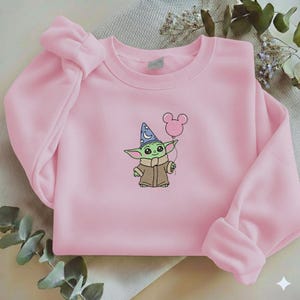 Op de afbeelding: Een lichtroze sweatshirt met een geborduurd cartoonfiguur met een tovenaars hoed en een roze ballon. Het figuur is groen met grote ogen en een bruine jas. De sweatshirt heeft lange mouwen.