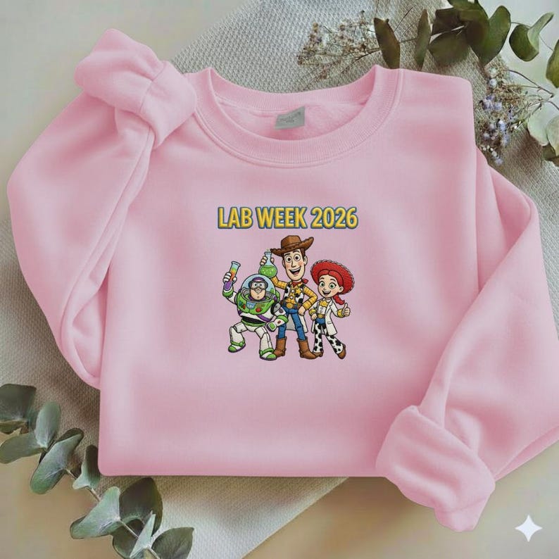 May include: A light pink sweatshirt featuring a graphic of three cartoon characters and the text "LAB WEEK 2026" in yellow. The characters include a space ranger, a cowboy, and a cowgirl. The sweatshirt has long sleeves and a crew neck.