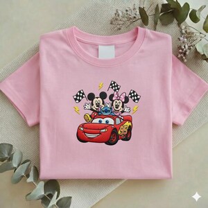May include: Pink short-sleeved t-shirt featuring an embroidered design of Mickey Mouse, Minnie Mouse, Stitch, and Lightning McQueen. The design includes checkered flags and the number 95. The shirt is made of a soft material.