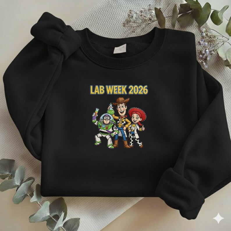 May include: Black crewneck sweatshirt featuring a graphic of Buzz Lightyear, Woody, and Jessie from Toy Story. The text "LAB WEEK 2026" is printed above the characters in yellow. The sweatshirt is made of a soft material.