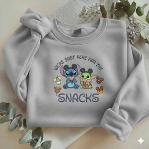 May include: A light gray sweatshirt featuring a graphic of Stitch and Grogu with snacks. The text "WE'RE JUST HERE FOR THE SNACKS" is embroidered below the characters. The sweatshirt has long sleeves and a crew neck.