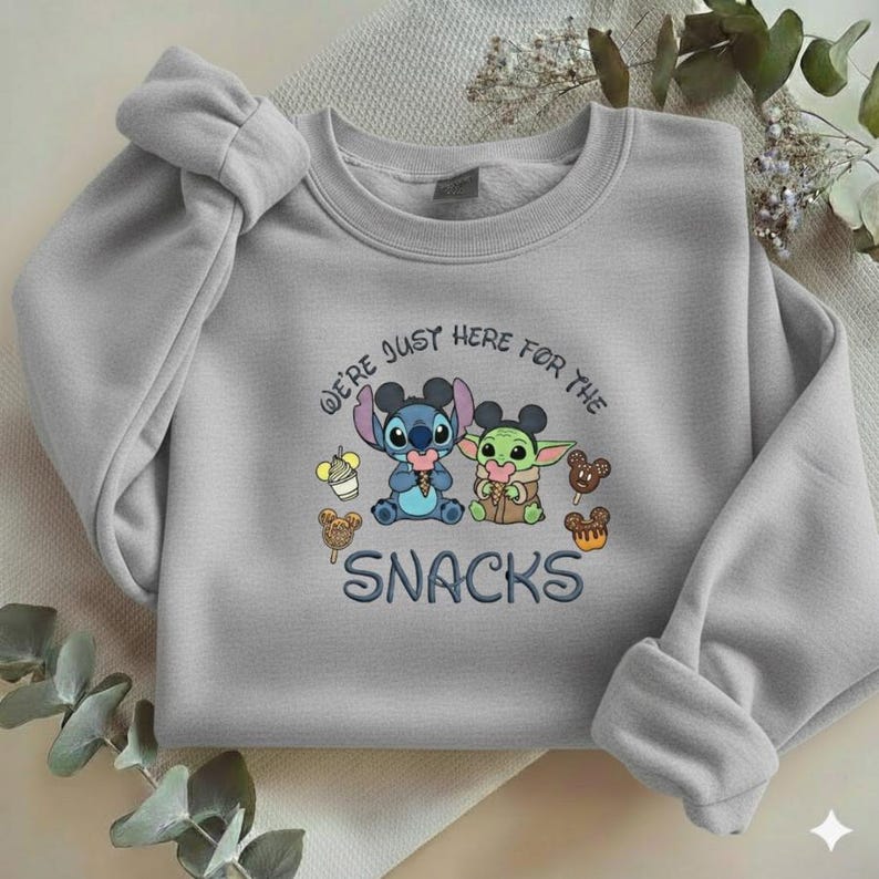 May include: A light gray sweatshirt featuring a graphic of Stitch and Grogu with snacks. The text "WE'RE JUST HERE FOR THE SNACKS" is embroidered below the characters. The sweatshirt has long sleeves and a crew neck.