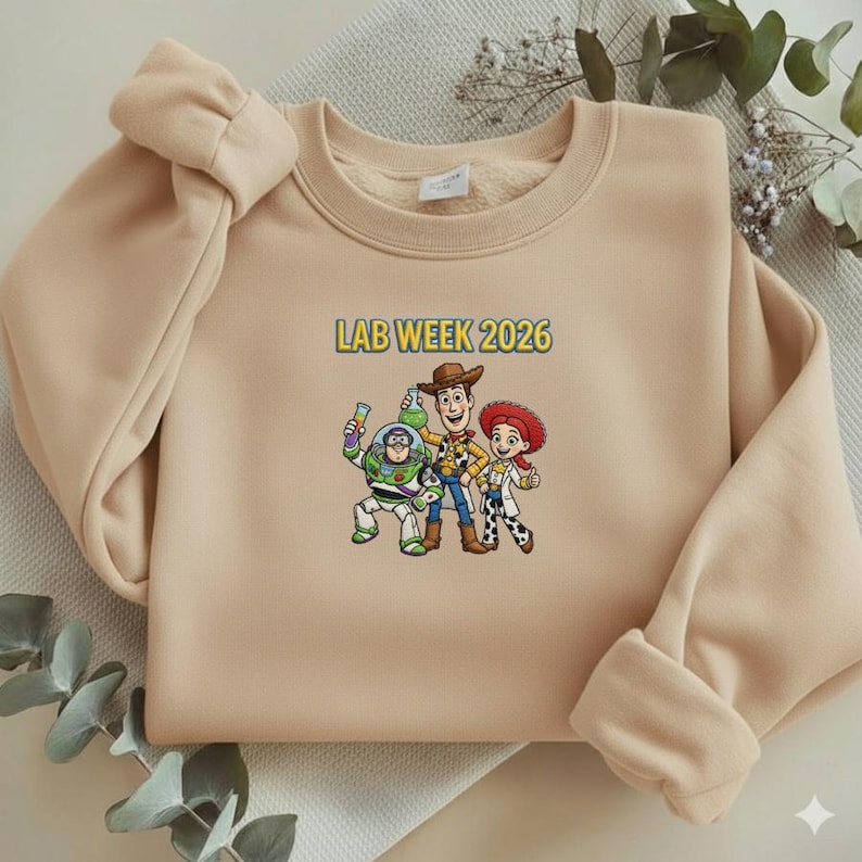 May include: A beige sweatshirt featuring a colorful graphic of three cartoon characters and the text "LAB WEEK 2026" in yellow and blue. The sweatshirt has long sleeves and a crew neck.
