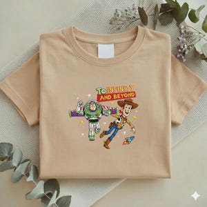 K&ouml;nnte beinhalten: Ein beiges T-Shirt mit einer bunten Grafik von Buzz Lightyear und Woody aus Toy Story, mit dem Text "To Infinity and Beyond". Das Design beinhaltet eine Rakete und Sterne. Das T-Shirt ist gefaltet.