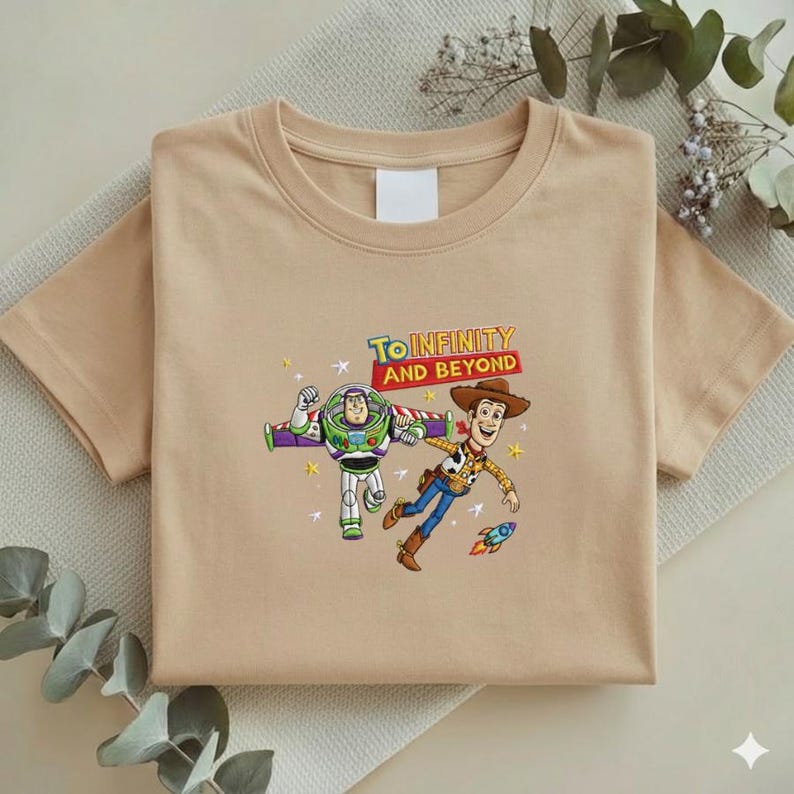K&ouml;nnte beinhalten: Ein beiges T-Shirt mit einer bunten Grafik von Buzz Lightyear und Woody aus Toy Story, mit dem Text "To Infinity and Beyond". Das Design beinhaltet eine Rakete und Sterne. Das T-Shirt ist gefaltet.