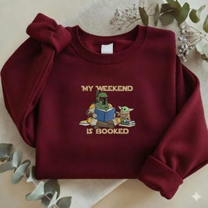 Pu&ograve; includere: Una felpa bordeaux con la scritta "MY WEEKEND IS BOOKED" e un'illustrazione di un personaggio che legge un libro con una piccola creatura verde. La felpa ha maniche lunghe e girocollo.