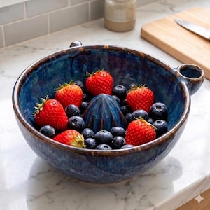 May include: A deep blue ceramic citrus juicer bowl with a central reamer, filled with fresh red strawberries and dark blue blueberries. The bowl features a glossy, mottled glaze finish and a small side handle, resting on a white marble surface.