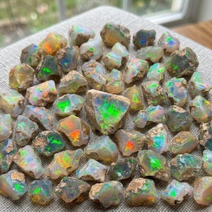 Peut inclure: Une collection de pierres d'opale brutes et non polies présentant une base laiteuse translucide avec des éclats vibrants de couleurs irisées, notamment du vert vif, de l'orange et du bleu, dispersés sur chaque pierre précieuse.