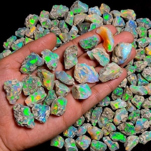 Peut inclure: Une collection de pierres précieuses d'opale brute, présentant une variété de couleurs, notamment le vert, le bleu, l'orange et le blanc. Les opales sont tenues dans une main, avec beaucoup d'autres dispersées en arrière-plan. Les pierres ont une texture naturelle rugueuse.
