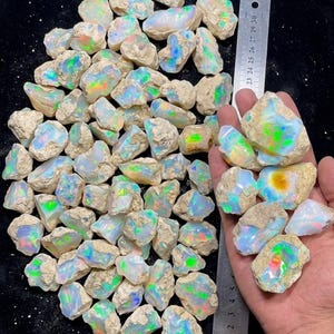 May include: A collection of raw, unpolished opal stones featuring iridescent flashes of green, blue, orange, and yellow against a milky white base. A metal ruler is visible, showing the stones range in size up to approximately 11 inches in length.