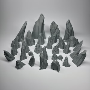 May include: A collection of grey, miniature rock formations of varying sizes and shapes. The rocks are arranged on a white surface, with some standing tall and others lying low. The overall aesthetic is minimalist and natural, suitable for tabletop gaming or model scenery.