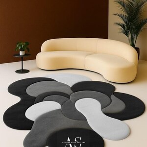 Puede incluir: Una alfombra moderna con un dise&ntilde;o abstracto &uacute;nico en tonos grises, negros y blancos. La alfombra presenta un patr&oacute;n floral con formas superpuestas. Las palabras "ARTISAN RUG GALLERY" est&aacute;n impresas en la alfombra.