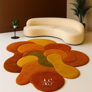 Puede incluir: Una alfombra abstracta en tonos naranja, amarillo y verde, con un dise&ntilde;o fluido. Un sof&aacute; curvo de color crema est&aacute; en el fondo. La alfombra est&aacute; sobre un suelo blanco. Las palabras "ARTISAN RUGS GALLERY" est&aacute;n impresas en la alfombra.