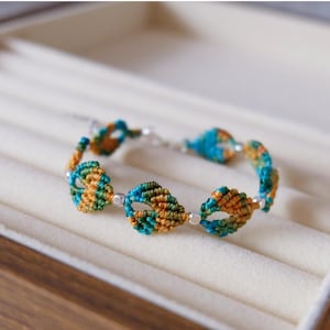Könnte beinhalten: Ein Makramee-Armband mit einer Reihe von blattförmigen Designs. Die Knoten des Armbands sind in Türkis, Orange und Grün gehalten, mit silberfarbenen Verbindungsgliedern und kleinen Perlen. Das Armband wird in einer hellen Schmuckschatulle präsentiert.