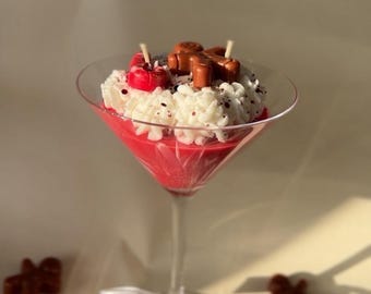 Może przedstawiać: Dekoracyjna świeca w kieliszku do martini. Świeca jest czerwona, zwieńczona białym woskiem, czerwoną wiśnią i piernikowym ludzikiem. Widoczne są dwa knoty. Biała wstążka jest zawiązana wokół nóżki kieliszka. Dodatkowi piernikowi ludziki są rozrzucone.