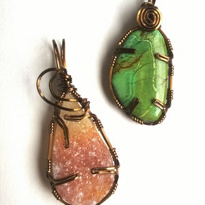 May include: Two wire-wrapped pendants with natural stone cabochons. The pendant on the left features a light orange and white stone with a rough, crystalline texture. The pendant on the right features a green stone with brown veins and a smooth, polished surface.