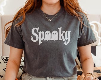 May include: Dark gray t-shirt with the word "Spooky" in white, featuring a ghost graphic. The shirt is a crew neck and has short sleeves. The model is wearing blue jeans and jewelry.