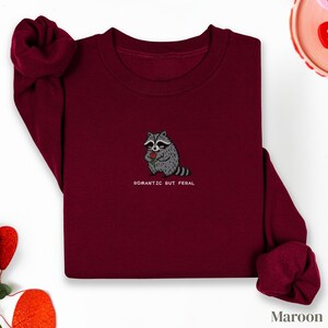 Può includere: Felpa bordeaux con un procione ricamato che tiene una rosa rossa. Il testo "ROMANTIC BUT FERAL" è sotto il procione. Le maniche sono arrotolate. La parola "Maroon" è in basso a destra.
