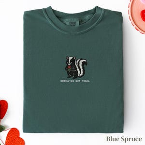 Può includere: Felpa girocollo blu abete con uno skunk ricamato che tiene una rosa rossa. Il testo "ROMANTIC BUT FERAL" è sotto. Prodotto da Comfort Colors.