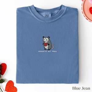 Può includere: T-shirt blu jeans con girocollo. Presenta un ricamo di un opossum che tiene un cuore rosso, con la scritta "ROMANTIC BUT FERAL" sotto. Il colletto mostra il marchio "COMFORT COLORS".