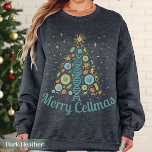 Peut inclure: Sweat-shirt gris foncé avec un motif d'arbre de Noël scientifique. L'arbre est composé de brins d'ADN, de cellules et d'étoiles, avec l'inscription "Merry Cellmas" en turquoise. Un vêtement de vacances festif et éducatif.