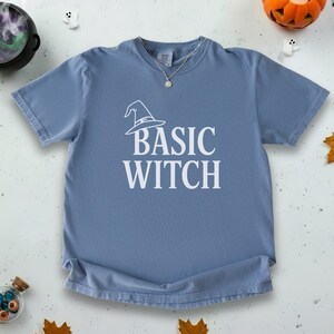 May include: A dusty blue t-shirt with the words "BASIC WITCH" in white block letters. A witch hat graphic is above the text. A silver necklace is visible at the neckline. The shirt is a casual, comfortable style.