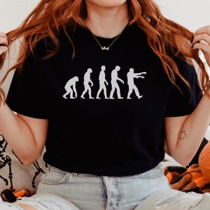 May include: Black t-shirt featuring a white graphic depicting the evolution of man into a zombie. The design shows a progression from a hunched figure to a walking zombie with outstretched arms. A fun and spooky design for Halloween.