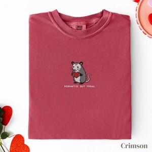 Può includere: Felpa color cremisi con girocollo. La maglietta presenta un opossum ricamato che tiene un cuore rosso, con la scritta "ROMANTIC BUT FERAL" sotto. Il marchio "COMFORT COLORS" è visibile sull'etichetta.