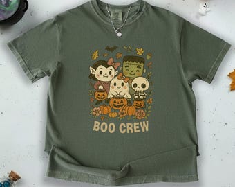 May include: Olive green t-shirt featuring a Halloween-themed graphic with cartoon characters, pumpkins, and the text "BOO CREW". The design includes a vampire, ghost, Frankenstein, and a skeleton. The shirt has a crew neck and short sleeves.