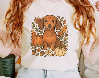 Op de afbeelding: Ivoor t-shirt met een bruine teckel illustratie, omringd door pompoenen, herfstbladeren, bloemen en eikels. Het ontwerp heeft een vintage, herfst esthetiek. Het shirt is gemaakt van een zacht materiaal.
