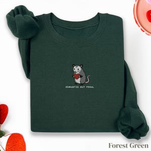 Può includere: Felpa verde foresta con un opossum ricamato che tiene un cuore rosso. Il testo "Romantic But Feral" è sotto. La maglia a maniche lunghe è realizzata in materiale morbido.