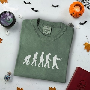May include: Sage green t-shirt with a white evolution of man to zombie graphic. The shirt has a crew neck and short sleeves. Halloween-themed decorations surround the shirt, including bats, a pumpkin, and a cauldron.