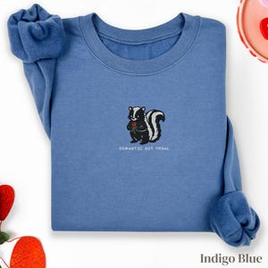 Può includere: Felpa blu indaco con un disegno di una puzzola dei cartoni animati che tiene una rosa rossa. La puzzola è bianca e nera con una coda soffice. Il testo "ROMANTIC BUT FERAL" è sotto. Le maniche sono arrotolate.
