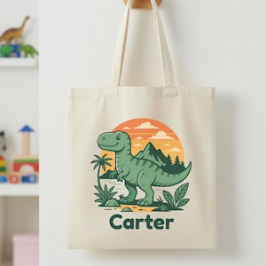 May include: Cream-colored tote bag featuring a green cartoon dinosaur with a mountain and sunset design. The name "Carter" is printed in black at the bottom. The bag has two cream-colored handles.