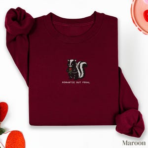 Può includere: Felpa bordeaux con ricamato un skunk che tiene una rosa rossa. Lo skunk è bianco e nero, con la scritta "ROMANTIC BUT FERAL" sotto. Le maniche sono arrotolate e la parola "Maroon" è in basso a destra.