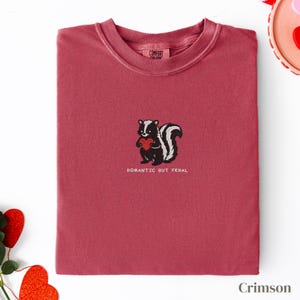 Può includere: Felpa color cremisi con girocollo. La maglietta presenta un ricamo di una puzzola che tiene un cuore rosso, con il testo "Romantic But Feral" sotto. La felpa è piegata ordinatamente.