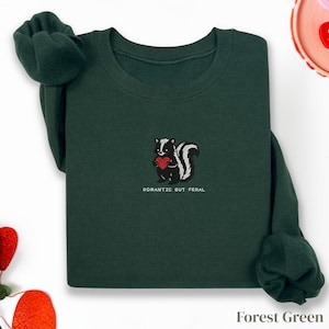 Può includere: Felpa verde foresta con una grafica di una puzzola che tiene un cuore rosso. Il testo "Romantic But Feral" è sotto la puzzola. Le maniche sono piegate. La felpa è su sfondo bianco.