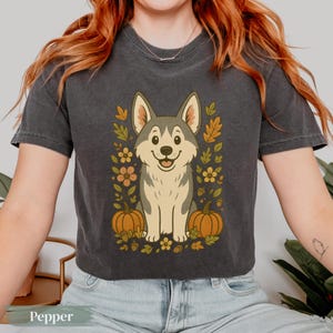 Puede incluir: Camiseta gris oscuro con un lobo de dibujos animados sonriendo, rodeado de hojas de otoño, calabazas y flores. La palabra "Pepper" está impresa en la parte inferior de la camiseta.