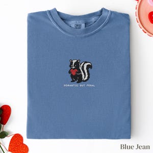 Può includere: T-shirt blu jeans con girocollo. Presenta una moffetta ricamata che tiene un cuore rosso, con la scritta "Romantic But Feral" sotto. L'etichetta del marchio Comfort Colors è visibile sul collo.