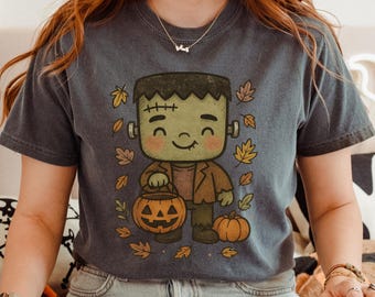 Puede incluir: Camiseta gris oscuro con un dibujo animado del monstruo de Frankenstein sosteniendo una linterna de calabaza. El diseño incluye hojas de otoño y una pequeña calabaza. El monstruo es verde con pelo castaño y una chaqueta marrón. Una divertida camiseta gráfica de Halloween.